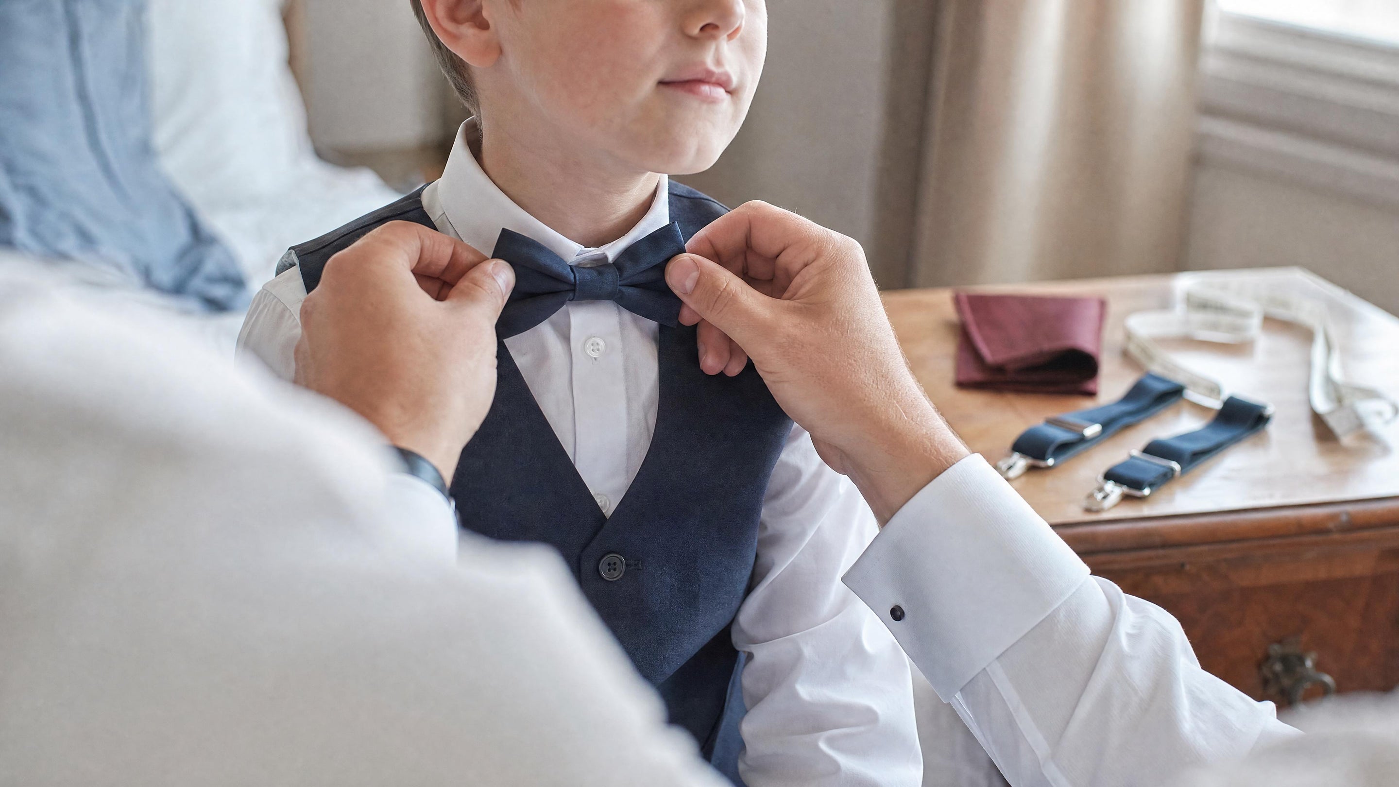 Un adulte ajuste un nœud papillon sur un enfant en chemise blanche, avec un mètre ruban visible sur une commode, dans une chambre lumineuse avant une cérémonie